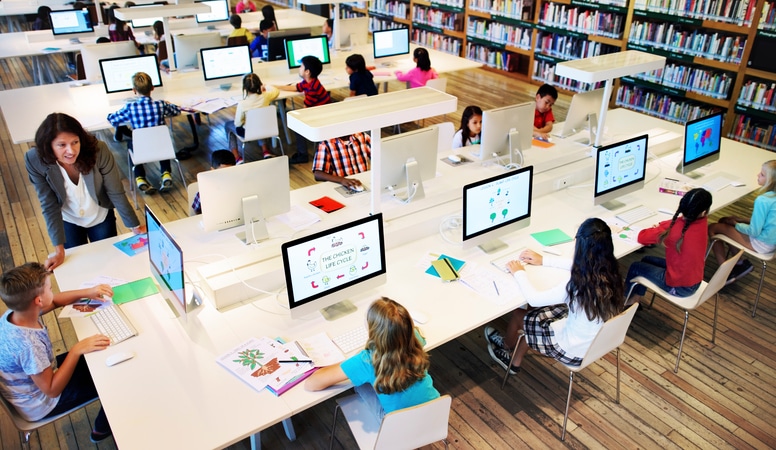 A stock photo shows a top-down angle of a library, where abut 10 young students sit around a table, each in front of their own computer.